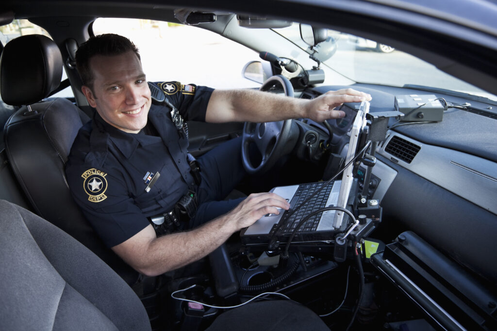 Police officer (20s) sitting in police car, with laptop computer.